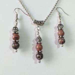 May include: A silver chain necklace with a silver pendant and two matching earrings. The pendant and earrings feature brown and black patterned beads.