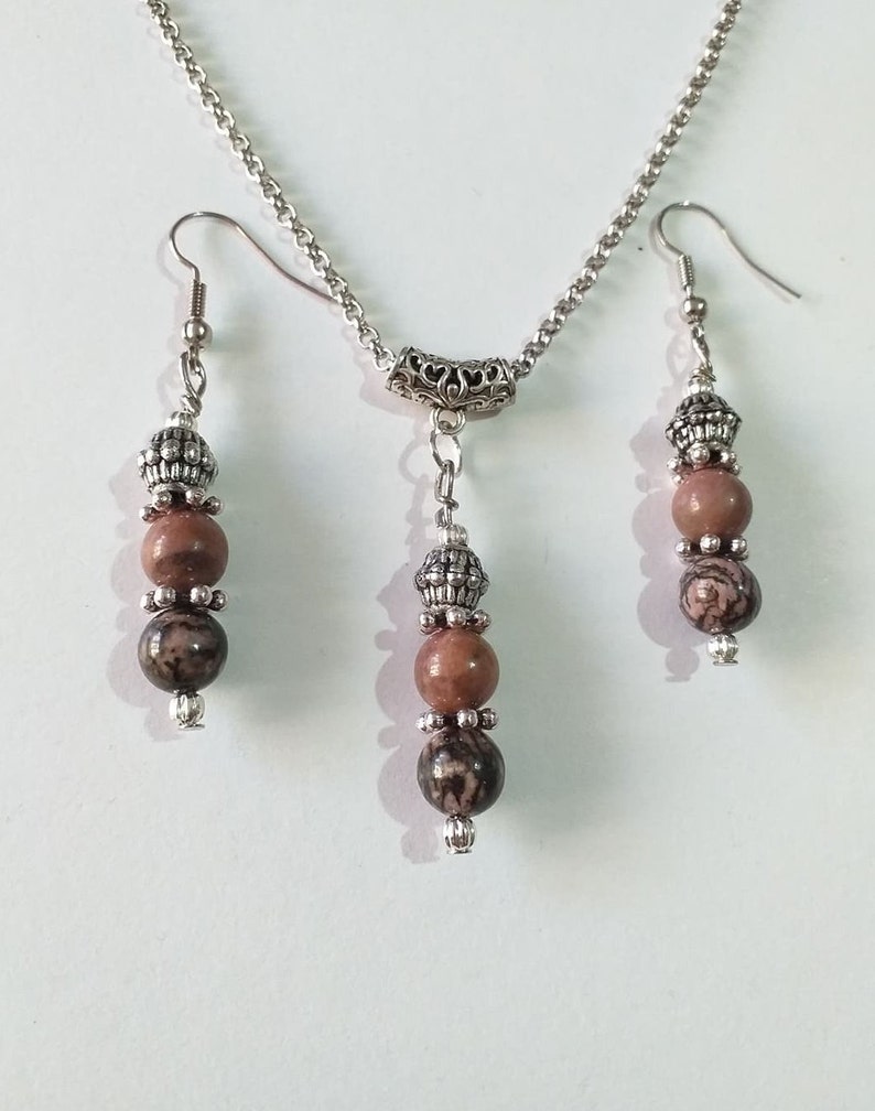 May include: A silver chain necklace with a silver pendant and two matching earrings. The pendant and earrings feature brown and black patterned beads.