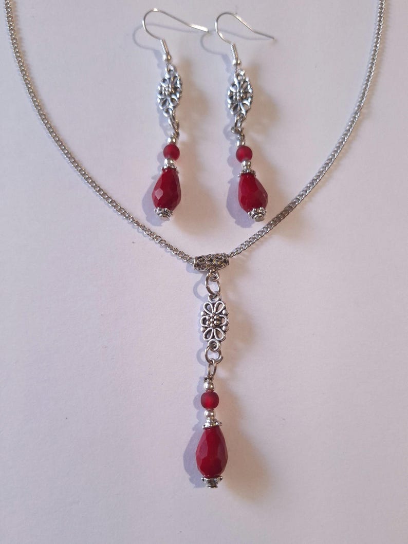 Puede incluir: Un collar de cadena de plata con un colgante de l&aacute;grima roja y un dije floral plateado. Aretes a juego con piedras de l&aacute;grima roja y dijes florales plateados.