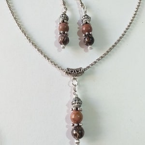 May include: A silver chain necklace with a pendant featuring three brown and black patterned beads. The necklace comes with a matching pair of earrings with three brown and black patterned beads.