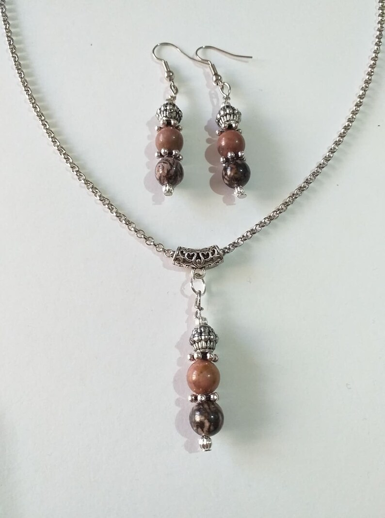 May include: A silver chain necklace with a pendant featuring three brown and black patterned beads. The necklace comes with a matching pair of earrings with three brown and black patterned beads.