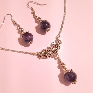 Puede incluir: Un collar de plata con un delicado colgante adornado con un dise&ntilde;o de hojas. El colgante est&aacute; adornado con una piedra preciosa redonda de color p&uacute;rpura oscuro. Pendientes a juego con piedras preciosas similares cuelgan de ganchos de plata.