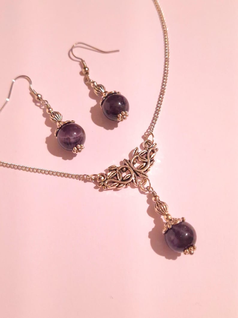 Puede incluir: Un collar de plata con un delicado colgante adornado con un dise&ntilde;o de hojas. El colgante est&aacute; adornado con una piedra preciosa redonda de color p&uacute;rpura oscuro. Pendientes a juego con piedras preciosas similares cuelgan de ganchos de plata.
