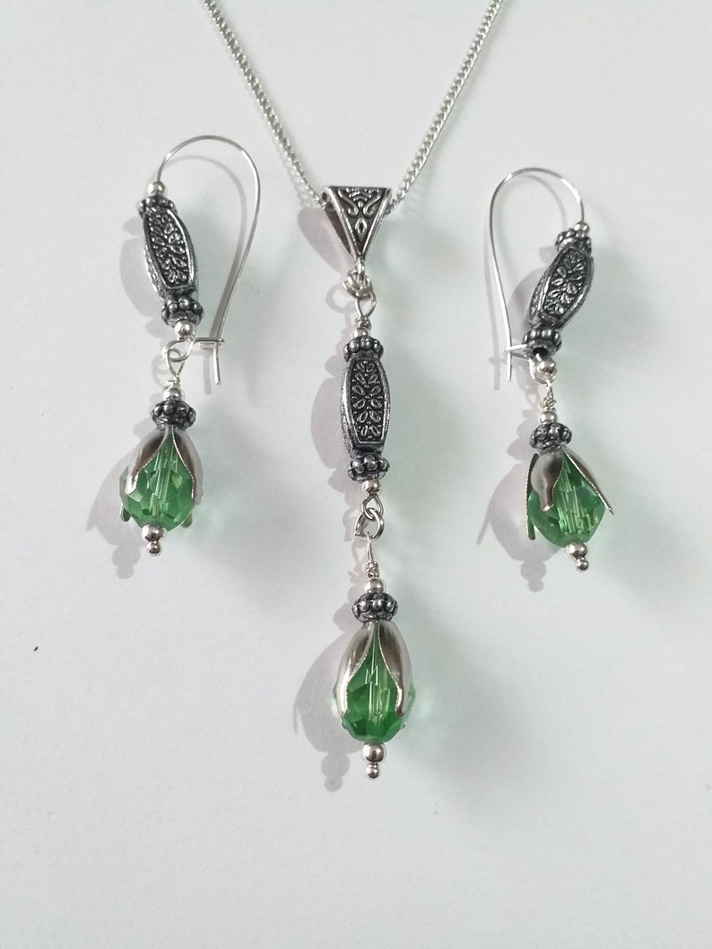 May include: A silver necklace and earrings set with green crystal teardrop pendants and ornate silver accents.