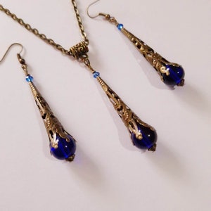 May include: A set of blue and gold jewelry featuring a necklace and earrings. The necklace has a delicate chain and a pendant with a blue glass bead and ornate gold filigree. The earrings are drop earrings with a similar design and blue glass beads.