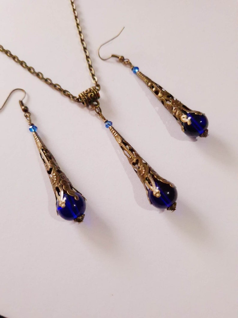 May include: A set of blue and gold jewelry featuring a necklace and earrings. The necklace has a delicate chain and a pendant with a blue glass bead and ornate gold filigree. The earrings are drop earrings with a similar design and blue glass beads.