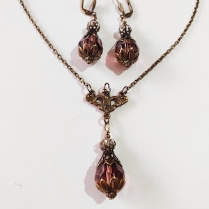 Op de afbeelding: Een set antieke sieraden met een delicate kettingketting met een hanger en bijpassende oorbellen. De hanger en oorbellen hebben elk een druppelvormig paars kristal met sierlijke metalen accenten.