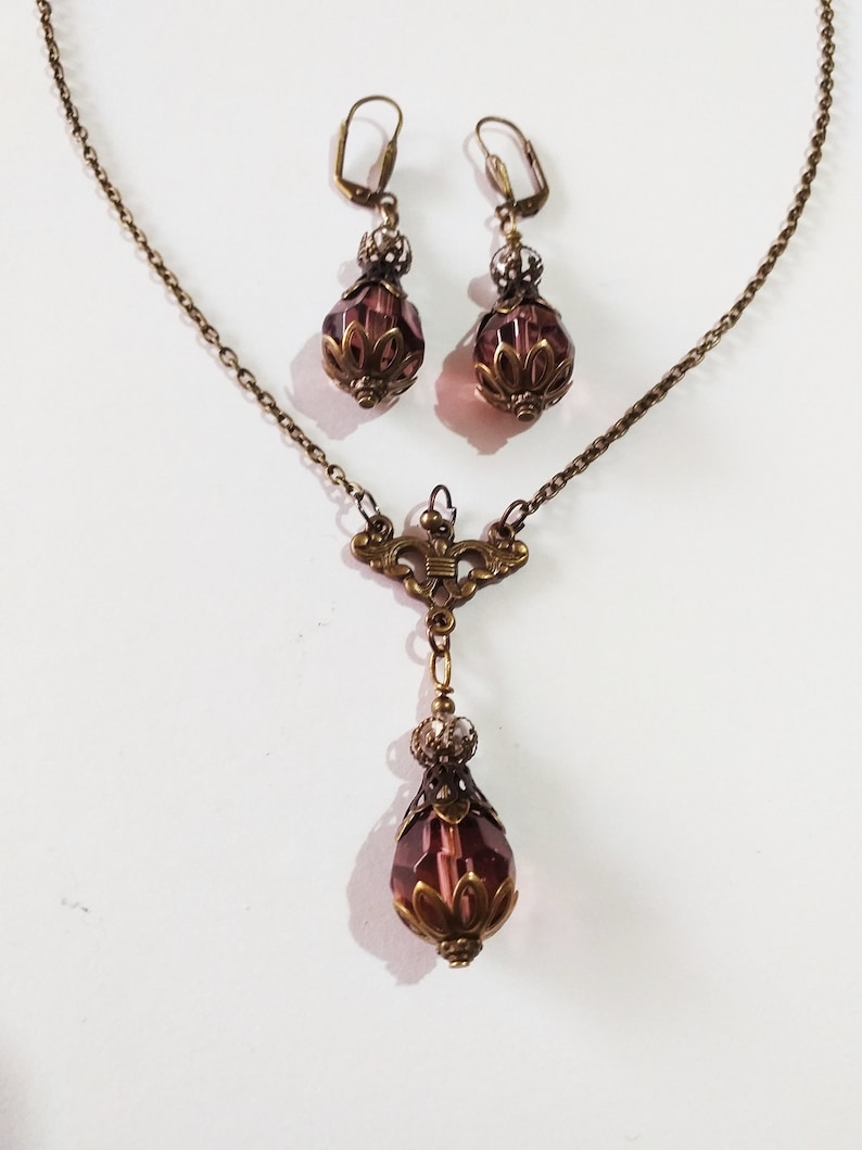 Op de afbeelding: Een set antieke sieraden met een delicate kettingketting met een hanger en bijpassende oorbellen. De hanger en oorbellen hebben elk een druppelvormig paars kristal met sierlijke metalen accenten.