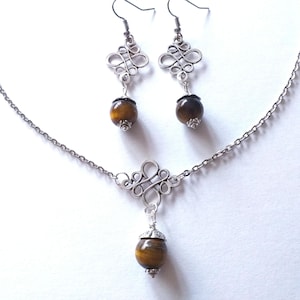 May include: A silver necklace and earrings set with a brown tiger's eye stone pendant and matching earrings. The necklace features a silver knot design and the earrings have a similar knot design.