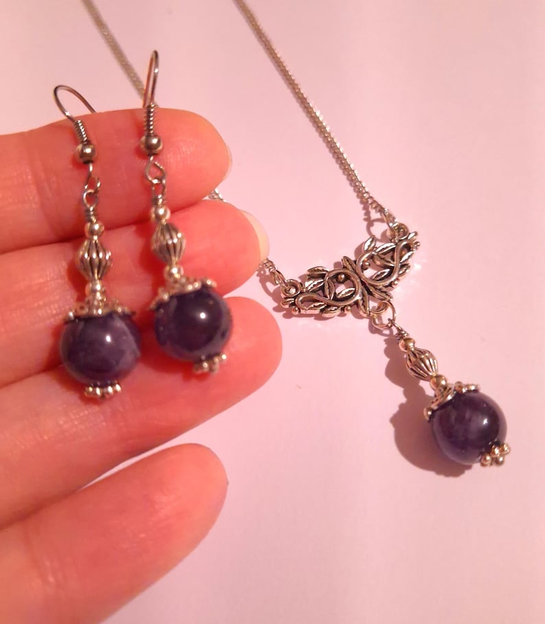Puede incluir: Un conjunto de collar y pendientes de plata con un colgante de piedra morada y pendientes a juego. El collar presenta un dise&ntilde;o delicado y adornado con una piedra colgante.