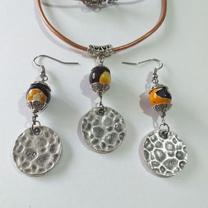 May include: A jewelry set featuring a brown leather cord necklace and matching earrings. The earrings and pendant have a decorative bead with yellow and brown accents, and a hammered silver disc. The necklace has a silver clasp.