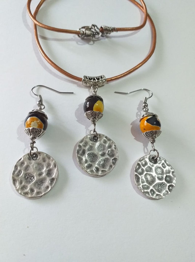May include: A jewelry set featuring a brown leather cord necklace and matching earrings. The earrings and pendant have a decorative bead with yellow and brown accents, and a hammered silver disc. The necklace has a silver clasp.