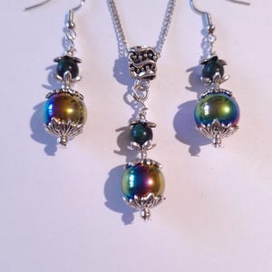 Può includere: Un set di collana e orecchini in argento con perline verdi e nere iridescenti. La collana ha una catena in argento e un ciondolo in argento con un design a spirale. Gli orecchini hanno ganci in argento e pendono dalle perline.