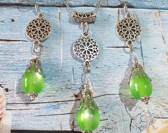 Puede incluir: Un collar y pendientes de plata con cuentas de vidrio verde y detalles de filigrana. El collar tiene un colgante con un diseño de filigrana redonda y dos cuentas de vidrio verde que cuelgan de él. Los pendientes tienen un diseño de filigrana redonda y una cuenta de vidrio verde que cuelga de cada uno.