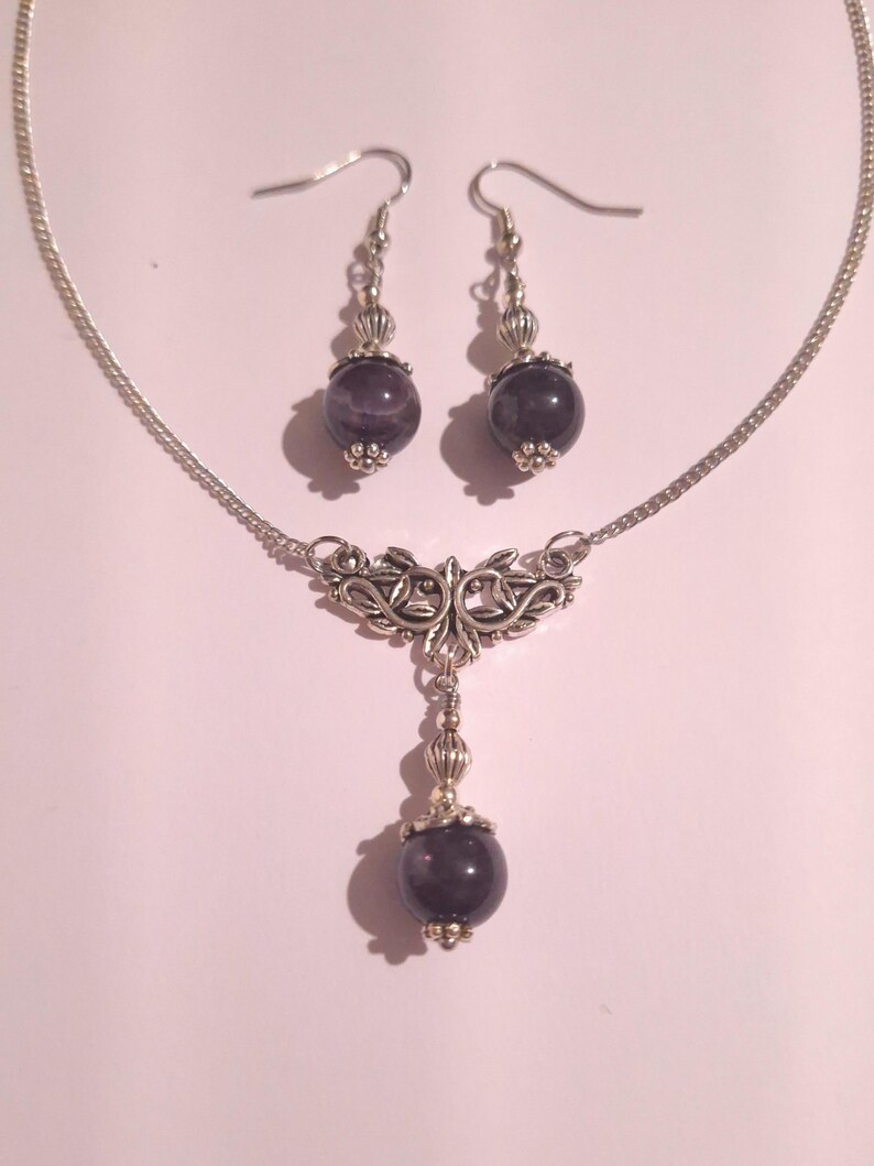 May include: A silver necklace and earrings set with a dark purple stone pendant and matching earrings. The necklace features a delicate silver chain and a decorative pendant with a floral design.
