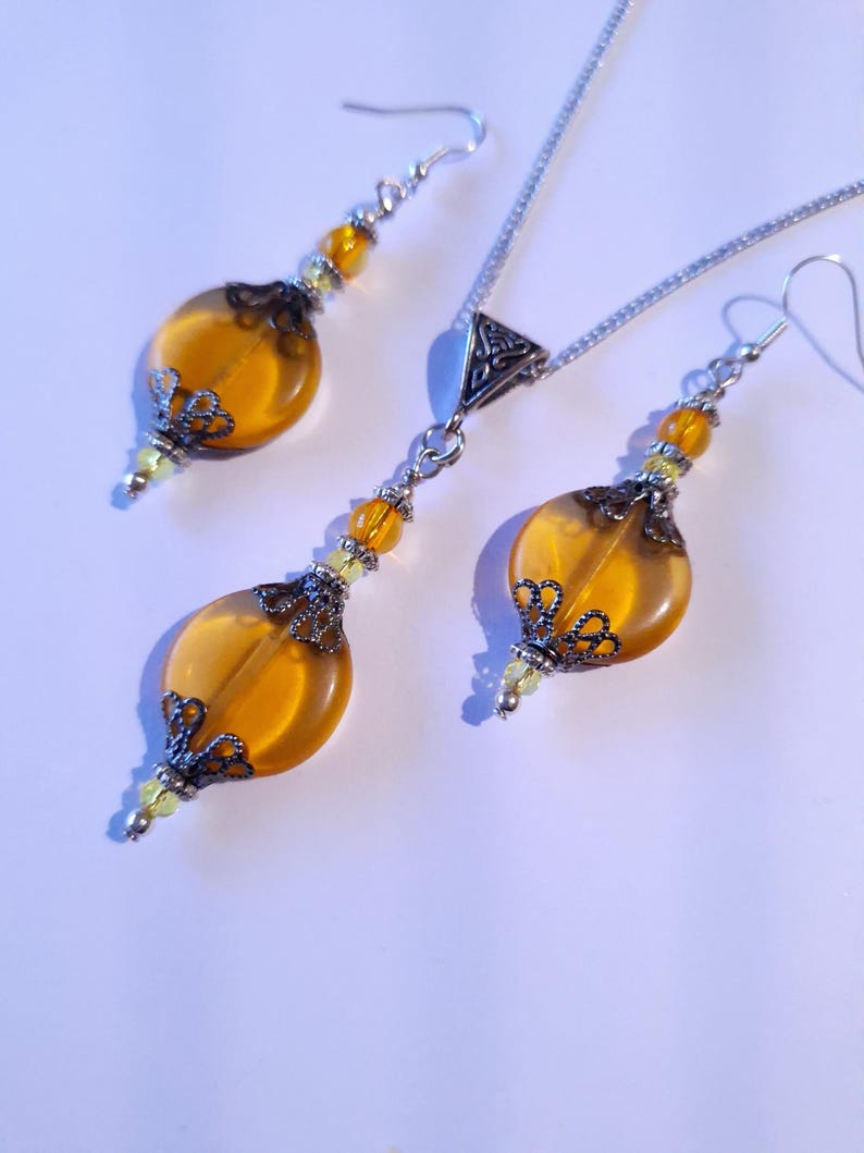 May include: A set of amber glass earrings and a necklace with silver chain and filigree accents. The earrings and necklace feature round amber glass beads with silver filigree accents.