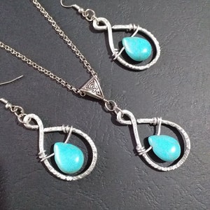 K&ouml;nnte beinhalten: Ein silbernes Halsketten- und Ohrring-Set mit t&uuml;rkisfarbenen Tr&auml;nensteinen. Die Halskette hat einen silbernen Anh&auml;nger mit einem komplizierten Design und die Ohrringe sind einfache Tr&auml;nchenformen.