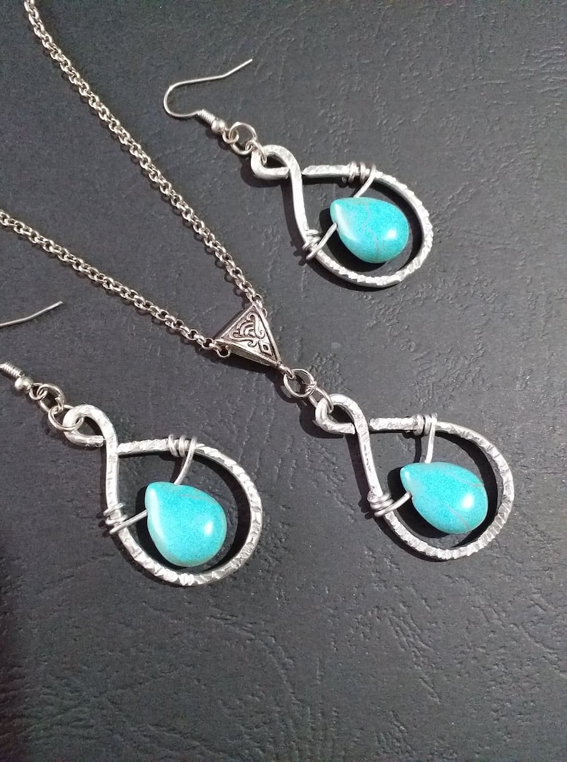 K&ouml;nnte beinhalten: Ein silbernes Halsketten- und Ohrring-Set mit t&uuml;rkisfarbenen Tr&auml;nensteinen. Die Halskette hat einen silbernen Anh&auml;nger mit einem komplizierten Design und die Ohrringe sind einfache Tr&auml;nchenformen.