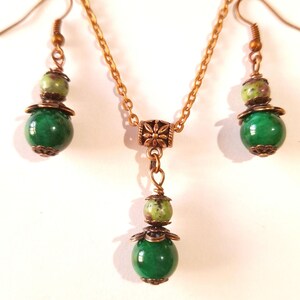 Può includere: Un set di collana e orecchini con pietre preziose verdi e marroni. La collana ha una catena dorata e un ciondolo con due pietre preziose verdi e marroni. Gli orecchini sono orecchini pendenti con una pietra preziosa verde e marrone.
