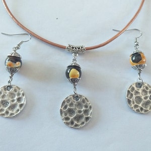 May include: A jewelry set featuring a brown leather choker necklace and matching earrings. The earrings and pendant have a black and yellow bead design, with a hammered silver disc. The jewelry set is a unique accessory.