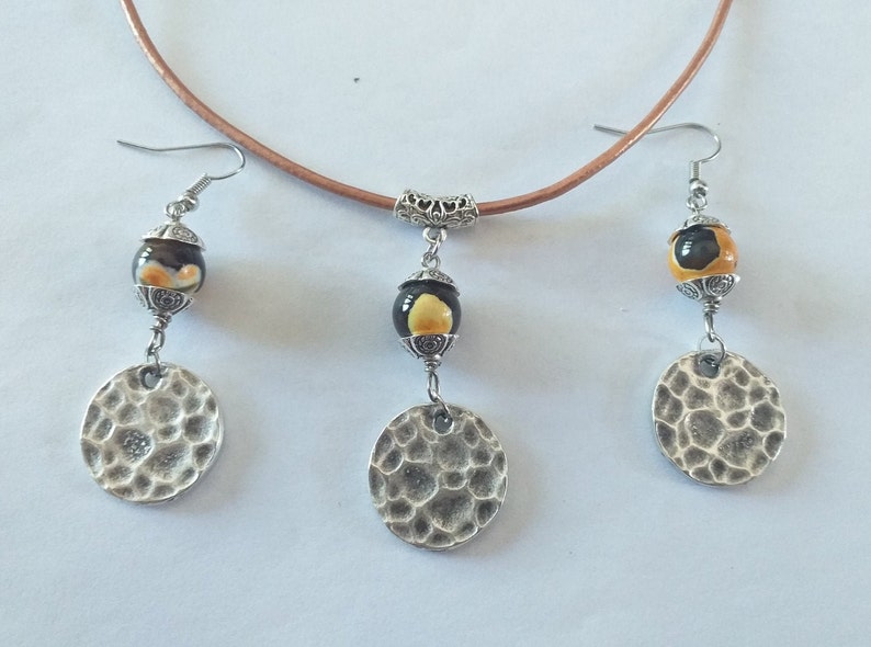 May include: A jewelry set featuring a brown leather choker necklace and matching earrings. The earrings and pendant have a black and yellow bead design, with a hammered silver disc. The jewelry set is a unique accessory.