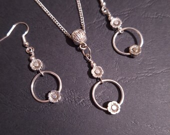 May include: A silver necklace and earrings set with a floral design. The necklace has a round pendant with a floral design and two smaller floral accents. The earrings have a similar design with a round hoop and a floral accent.