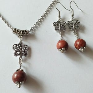 May include: A silver necklace and earrings set with a red stone pendant and butterfly charms. The necklace has a silver chain and a silver butterfly charm hanging from the red stone pendant. The earrings have silver butterfly charms hanging from red stone beads.