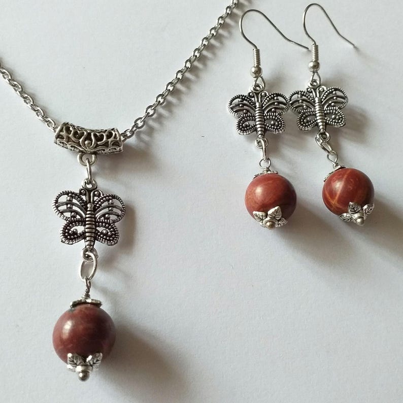 May include: A silver necklace and earrings set with a red stone pendant and butterfly charms. The necklace has a silver chain and a silver butterfly charm hanging from the red stone pendant. The earrings have silver butterfly charms hanging from red stone beads.