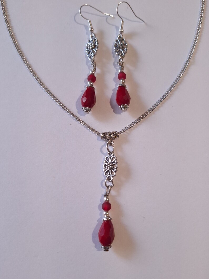 Puede incluir: Un collar de cadena de plata con un colgante de l&aacute;grima roja y un dije floral plateado. Aretes a juego con colgantes de l&aacute;grima roja y dijes florales plateados.