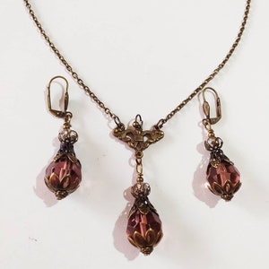Op de afbeelding: Een set sieraden met een delicate gouden ketting met een hanger en bijpassende oorbellen. De hanger en oorbellen hebben een traanvorm met een facetgeslepen paarse kristal en sierlijke gouden accenten.