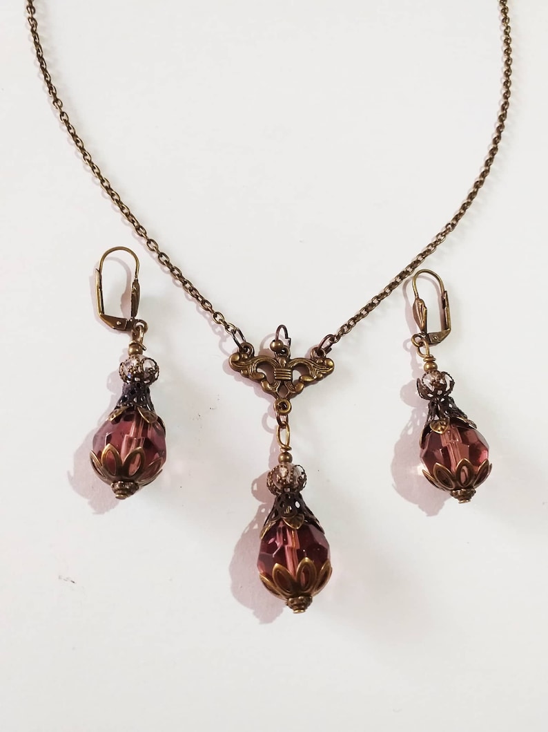Op de afbeelding: Een set sieraden met een delicate gouden ketting met een hanger en bijpassende oorbellen. De hanger en oorbellen hebben een traanvorm met een facetgeslepen paarse kristal en sierlijke gouden accenten.