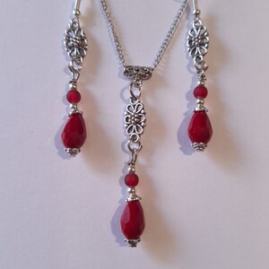 Puede incluir: Un collar y pendientes de plata con cuentas de vidrio rojas en forma de l&aacute;grima y detalles florales plateados.