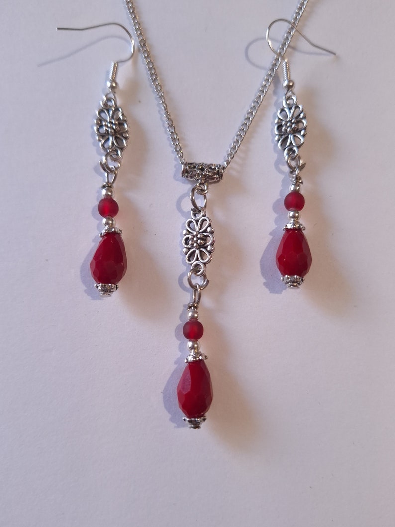 Puede incluir: Un collar y pendientes de plata con cuentas de vidrio rojas en forma de l&aacute;grima y detalles florales plateados.