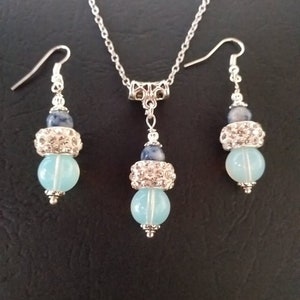 May include: A silver necklace and earrings set with blue and light blue gemstone beads. The necklace has a silver chain and a silver pendant with a blue gemstone bead and a light blue gemstone bead. The earrings have silver hooks and a blue gemstone bead and a light blue gemstone bead.