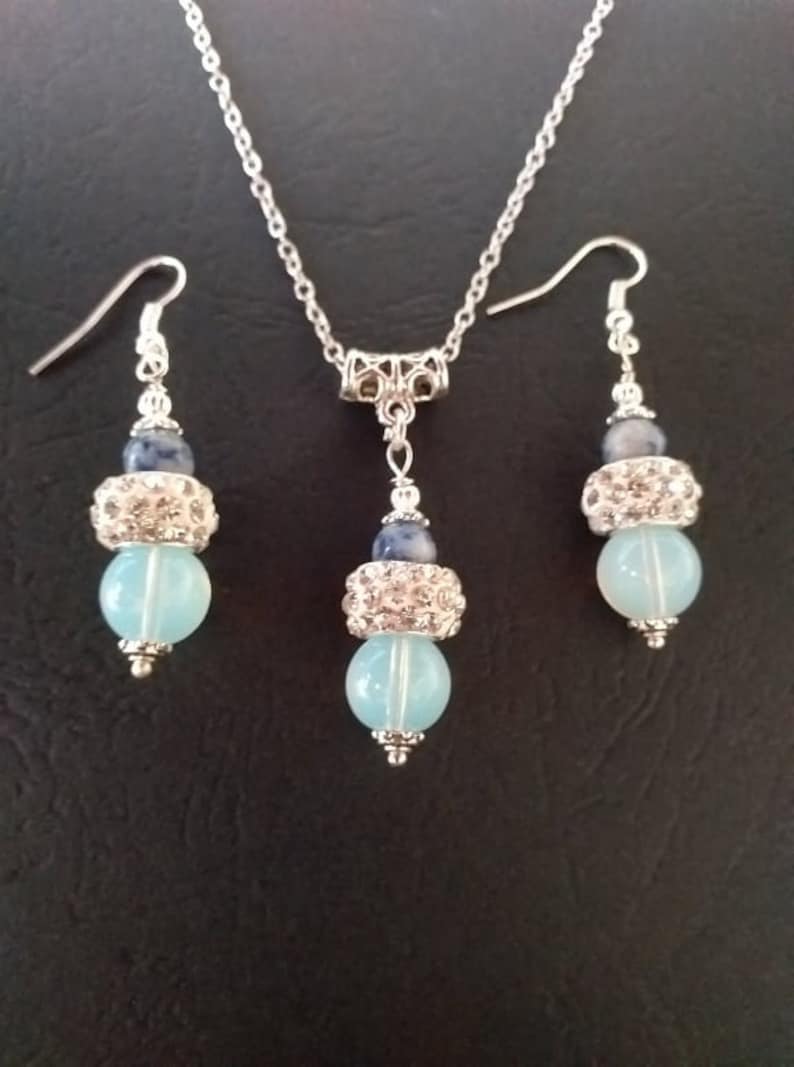 May include: A silver necklace and earrings set with blue and light blue gemstone beads. The necklace has a silver chain and a silver pendant with a blue gemstone bead and a light blue gemstone bead. The earrings have silver hooks and a blue gemstone bead and a light blue gemstone bead.