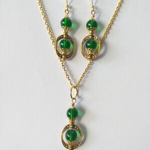 May include: A gold necklace and earrings set with green glass beads and gold accents. The necklace has a pendant with a circle design and two green beads. The earrings have a similar design with one green bead.