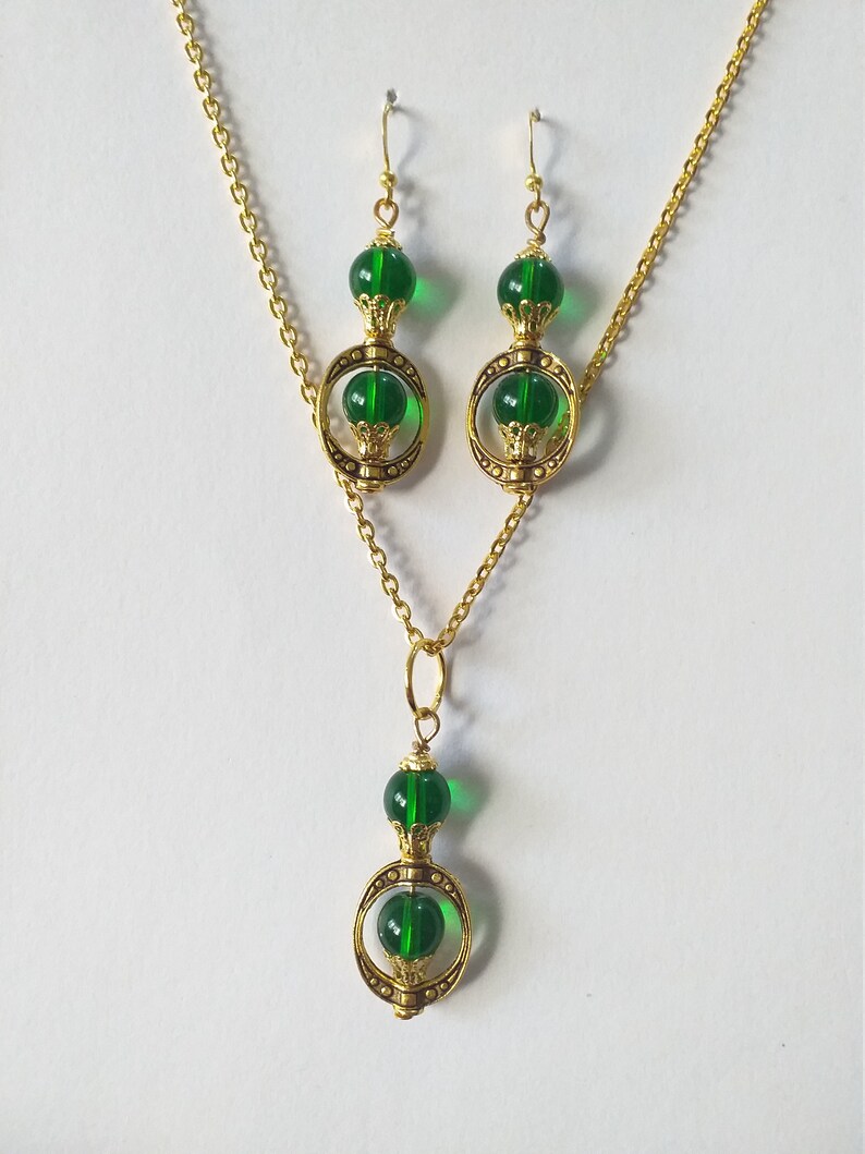 May include: A gold necklace and earrings set with green glass beads and gold accents. The necklace has a pendant with a circle design and two green beads. The earrings have a similar design with one green bead.