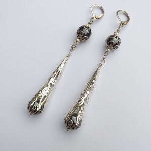 May include: A pair of silver dangle earrings with black beads and intricate filigree designs. The earrings have a teardrop shape and are suspended from a silver hook.