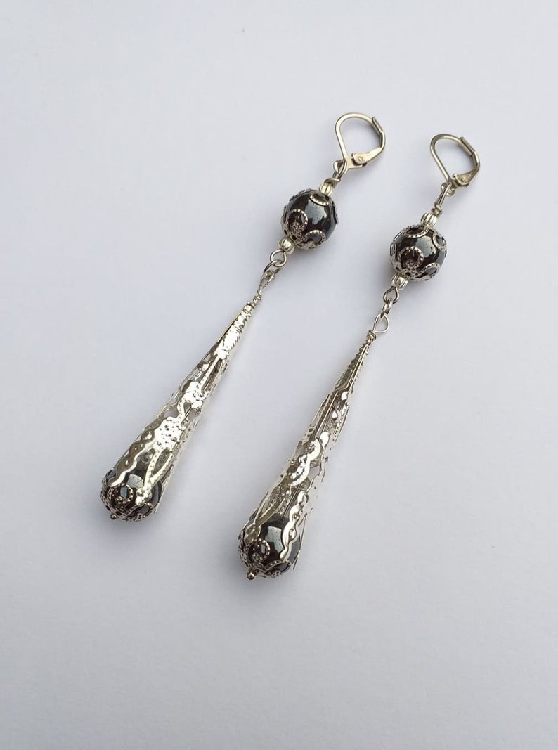 May include: A pair of silver dangle earrings with black beads and intricate filigree designs. The earrings have a teardrop shape and are suspended from a silver hook.