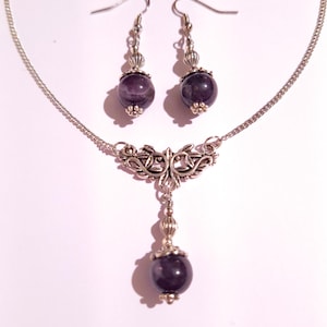 May include: A silver necklace with a pendant featuring a silver filigree design and a purple amethyst stone. Matching earrings with purple amethyst stones dangle from silver hooks.