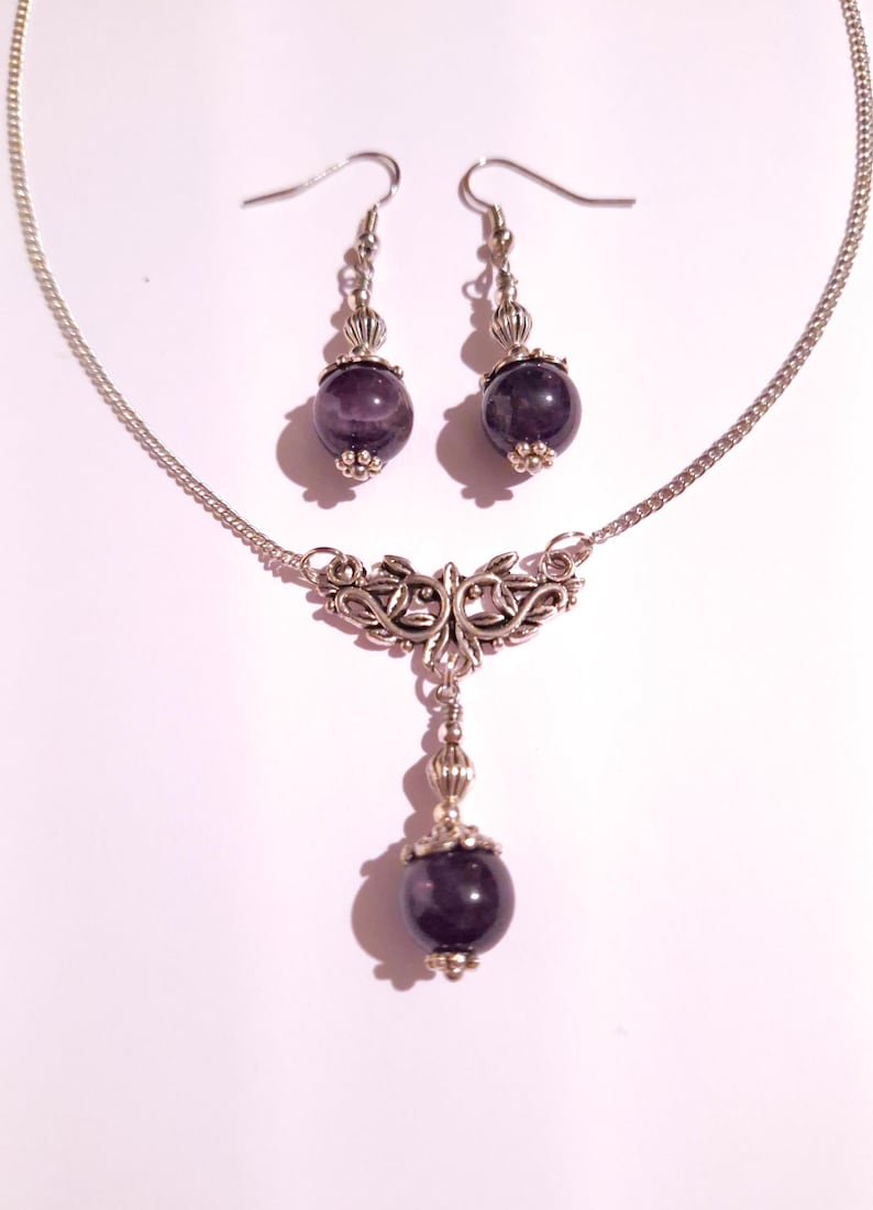 May include: A silver necklace with a pendant featuring a silver filigree design and a purple amethyst stone. Matching earrings with purple amethyst stones dangle from silver hooks.