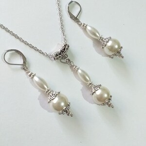 May include: A silver necklace and earrings set with white pearl accents. The necklace features a silver chain with a decorative pendant and a single pearl drop. The earrings are silver with a single pearl drop.