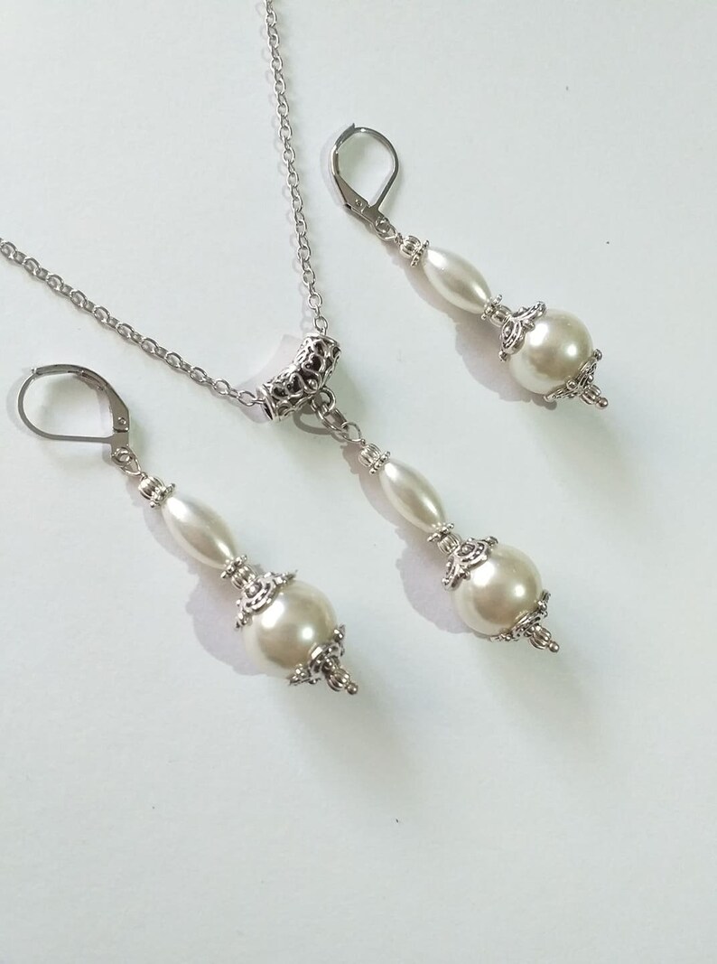 May include: A silver necklace and earrings set with white pearl accents. The necklace features a silver chain with a decorative pendant and a single pearl drop. The earrings are silver with a single pearl drop.