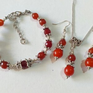Puede incluir: Un conjunto de joyas de piedra de cornalina roja con detalles plateados. El conjunto incluye un brazalete, un collar y un par de pendientes.