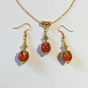 K&ouml;nnte beinhalten: Ein goldenes Halsketten- und Ohrring-Set mit Karneol-Edelsteinkugeln. Die Halskette hat eine goldene Kette und einen Anh&auml;nger mit einer Karneol-Kugel. Die Ohrringe haben goldene Haken und Karneol-Kugeln.