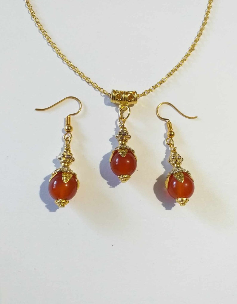 K&ouml;nnte beinhalten: Ein goldenes Halsketten- und Ohrring-Set mit Karneol-Edelsteinkugeln. Die Halskette hat eine goldene Kette und einen Anh&auml;nger mit einer Karneol-Kugel. Die Ohrringe haben goldene Haken und Karneol-Kugeln.