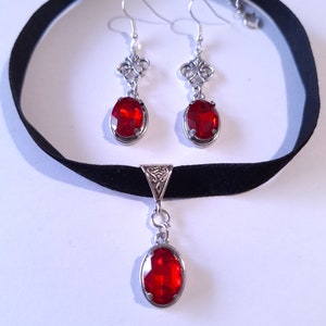 May include: A black velvet choker necklace with a silver pendant featuring a red oval-shaped gemstone. Matching earrings with silver filigree and red oval-shaped gemstones.
