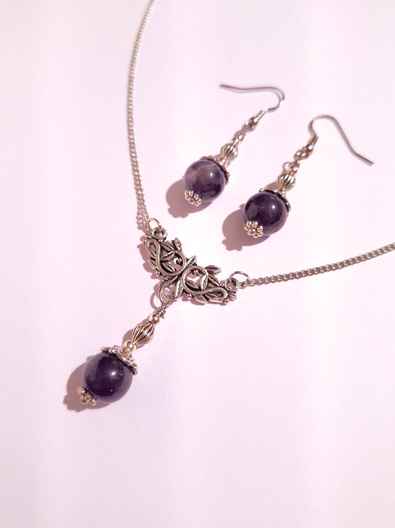Puede incluir: Un collar de plata con un delicado colgante floral y una piedra preciosa de amatista colgante. Pendientes de amatista a juego con ganchos de plata.
