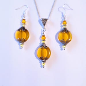 May include: A set of amber glass and silver tone jewelry. The necklace and earrings feature a round amber glass bead with a silver tone filigree design. The necklace has a silver tone chain.