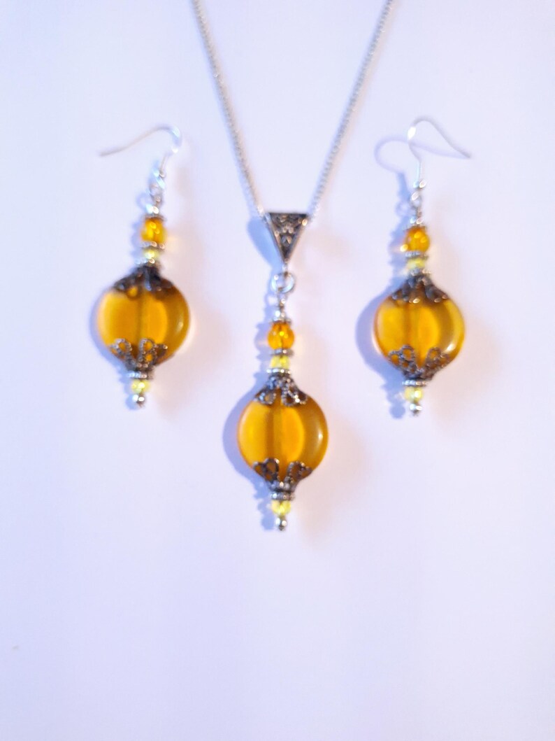 May include: A set of amber glass and silver tone jewelry. The necklace and earrings feature a round amber glass bead with a silver tone filigree design. The necklace has a silver tone chain.